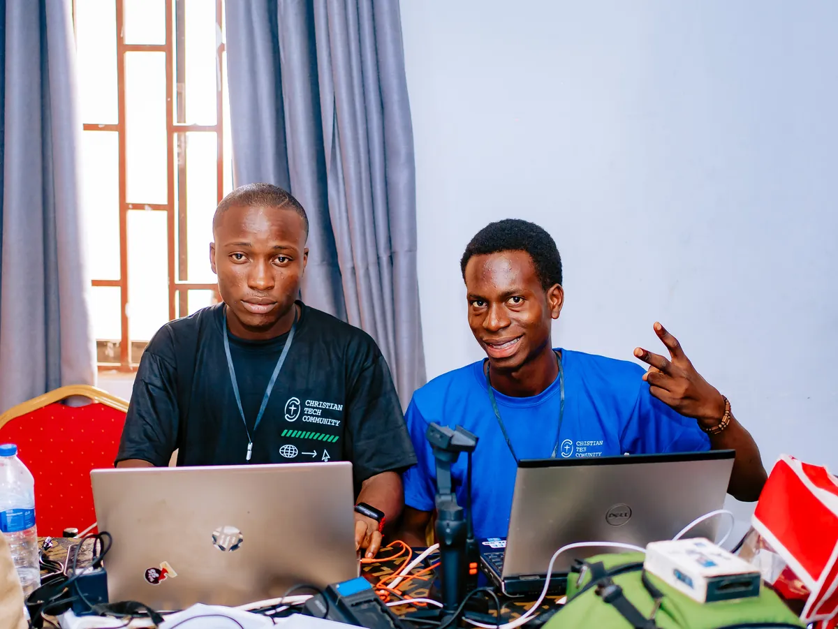 Two team members with lanyards sit behind laptops and cables at a tech desk at CTC 2.0, one making a peace sign.